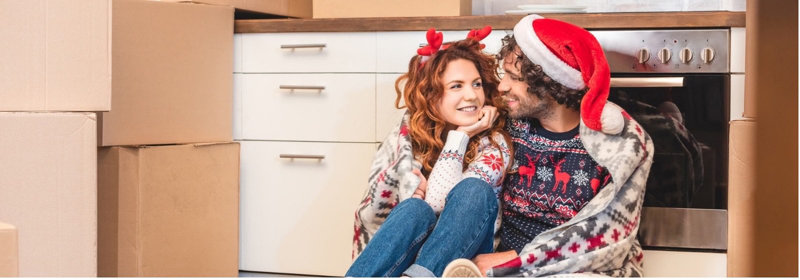 A man and woman sat down on the floor of a kitchen wearing Christmas hats.